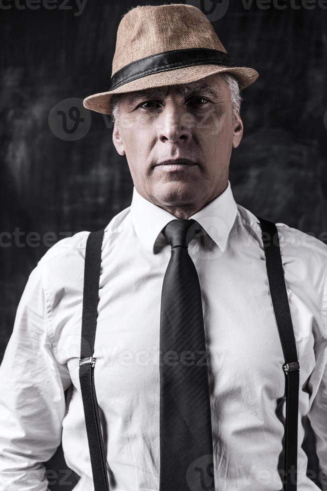 I am a boss hear Portrait of confident senior man in hat and suspenders looking at camera while standing against dark background photo