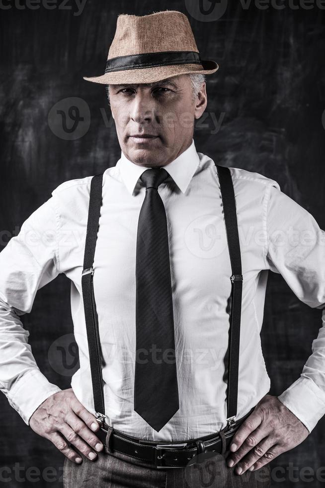 I am a boss Confident senior man in hat and suspenders looking at camera and holding hands on hip while standing against dark background photo