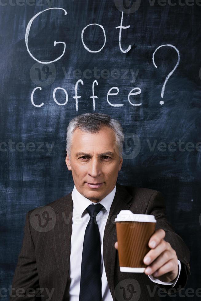 Take a break Confident senior man in formalwear stretching out coffee cup and smiling while standing against blackboard with chalk drawing on it photo