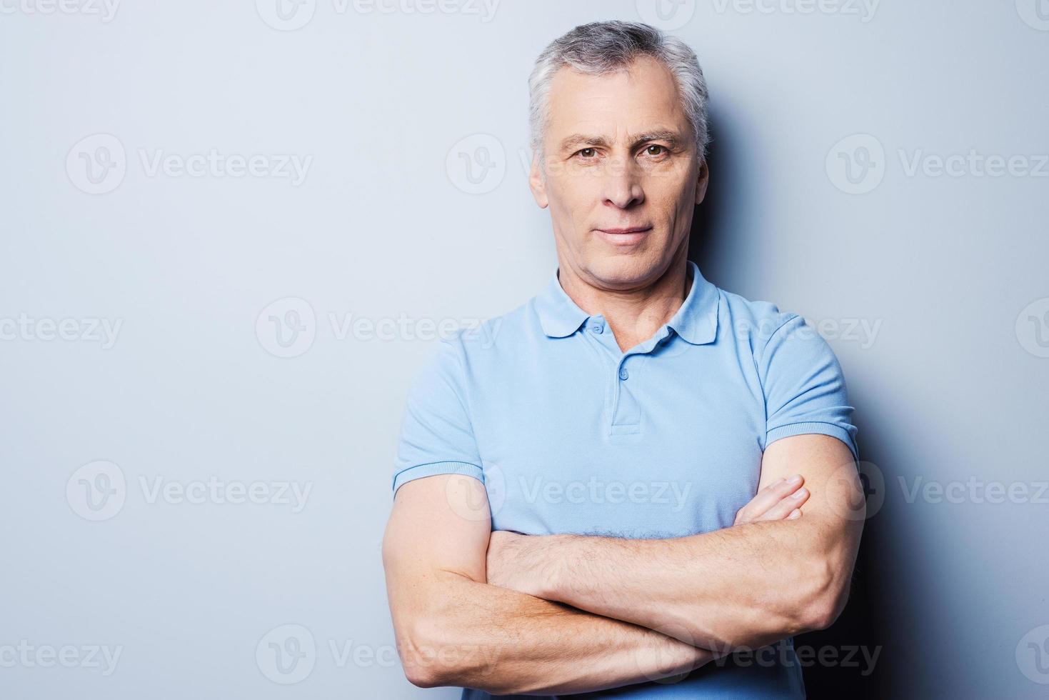 Confident mature man. Portrait of confident senior man in T-shirt looking at camera and smiling while keeping arms crossed and standing against grey background photo