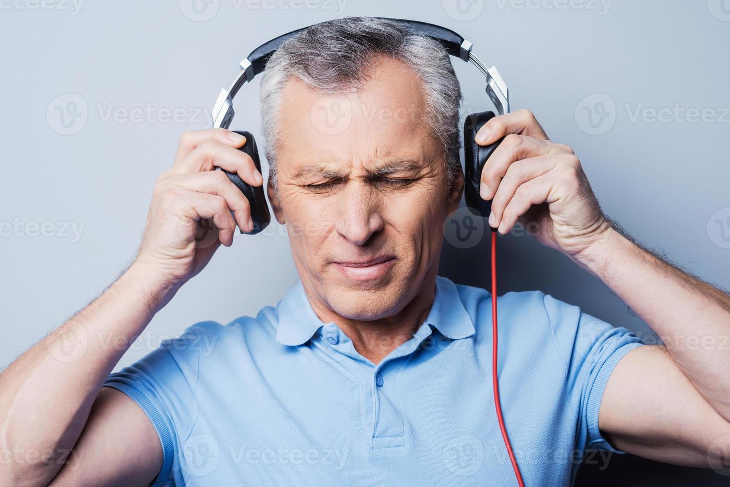 Such music is not for me. Portrait of frustrated senior man in headphones listening to music keeping eyes closed while standing against grey background photo