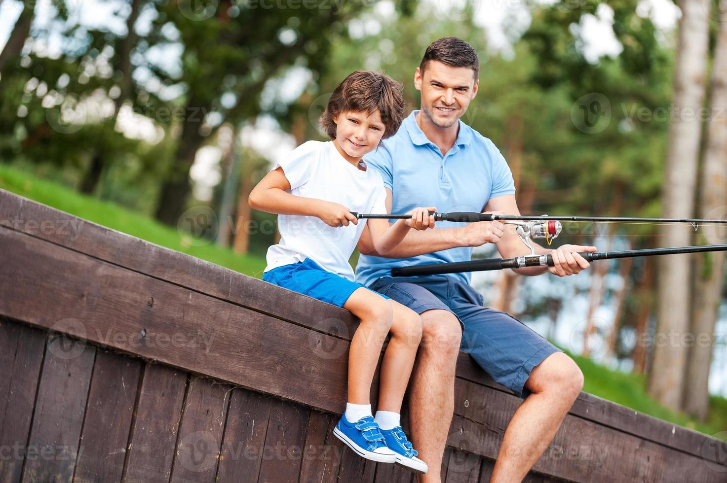 nos encanta pescar juntos. alegre padre e hijo pescando juntos y sonriendo mientras están sentados en el muelle foto