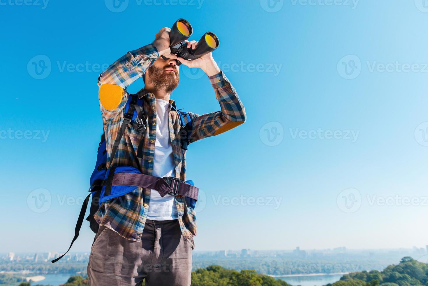 admirando la majestuosa vista. apuesto joven barbudo llevando mochila y mirando a través de binoculares foto