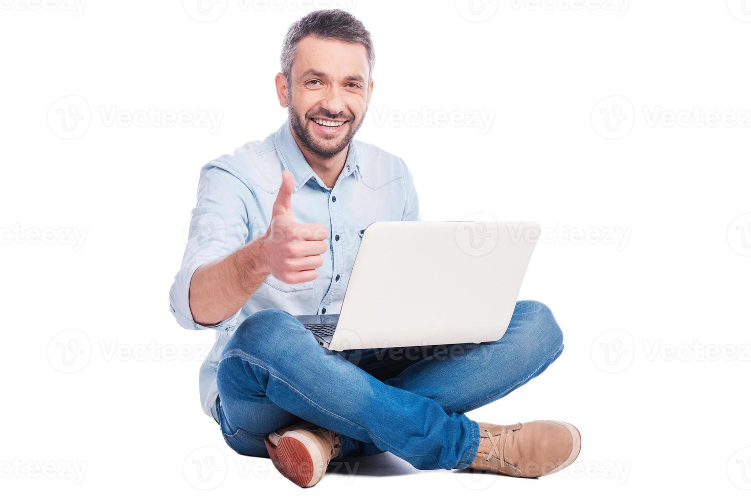 Technologies become easier Handsome young man in casual wear sitting on the floor with laptop and showing his thumb up while being isolated on white background photo