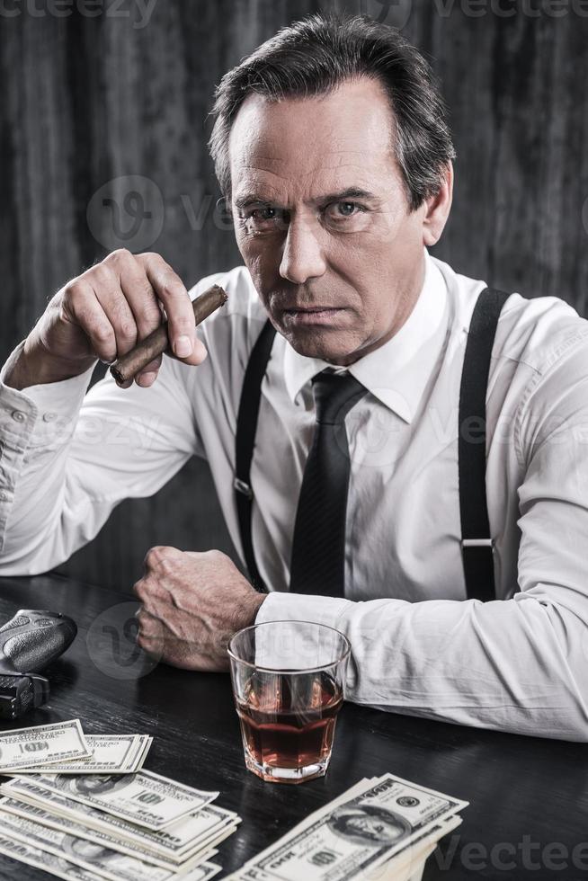 What do you want Top view of serious senior man in shirt and suspenders sitting at the table and holding cigar while lots of money laying near him photo