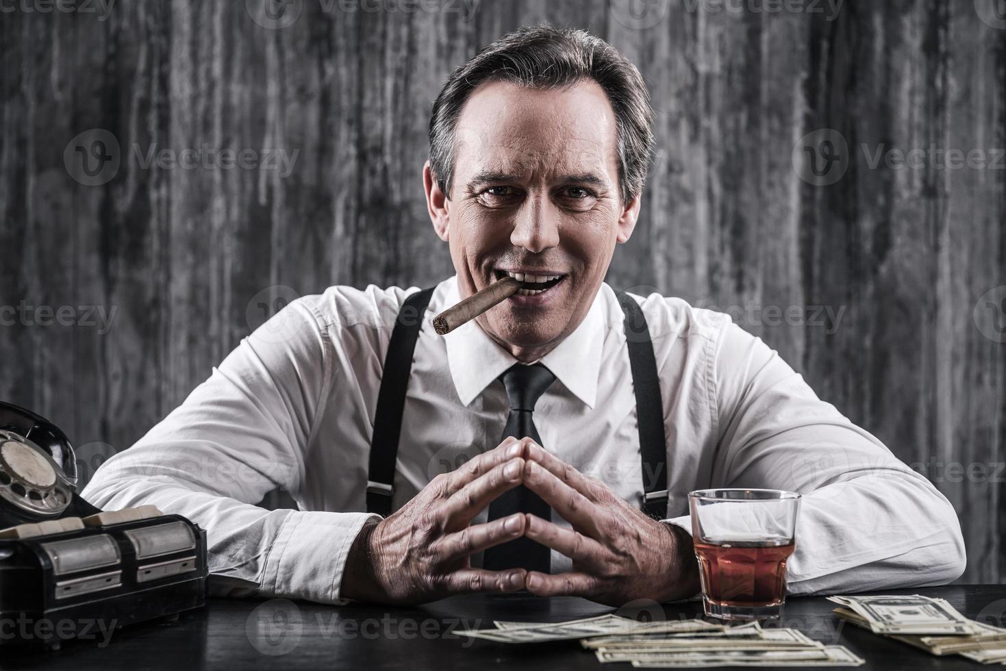 Power is in his hands. Smiling senior man in shirt and suspenders sitting at the table and smoking cigar while lots of money laying near him photo