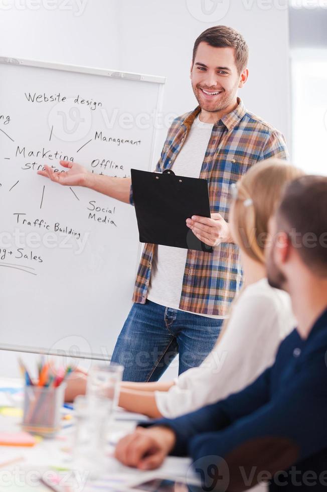 Confident in his speech. Business people in smart casual wear sitting together at the table while confident young man standing near whiteboard and pointing it with smile photo