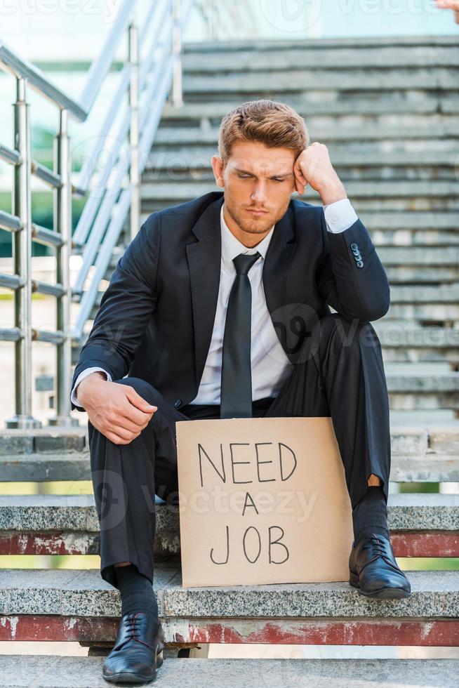 Need a job. Depressed young man in formalwear holding poster with job text message while sitting on the staircase photo