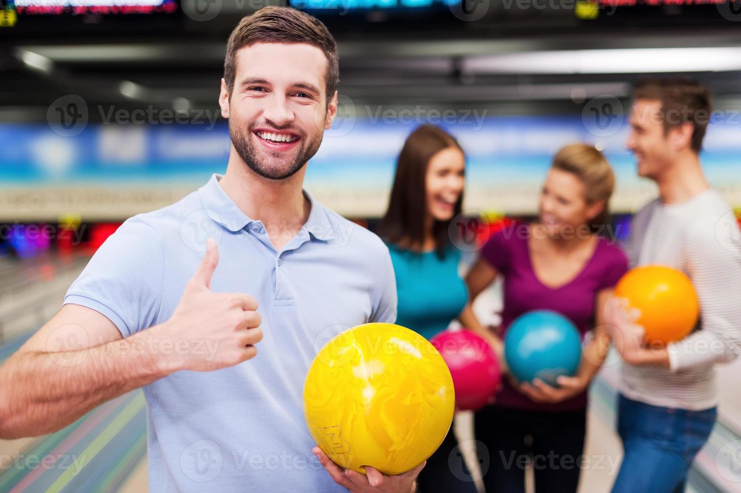 Great game Handsome young men holding a bowling ball  and thumb up while three people communicating against bowling alleys photo
