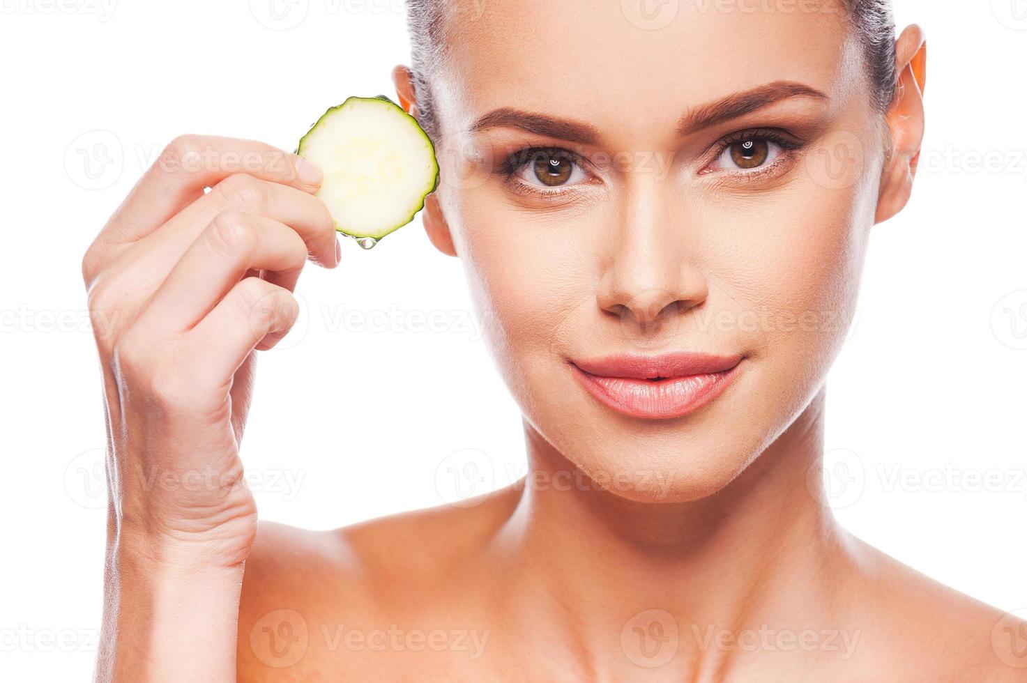 Being fresh is simple deal. Close-up of beautiful young shirtless woman holding slice of cucumber and looking at camera while standing against white background photo