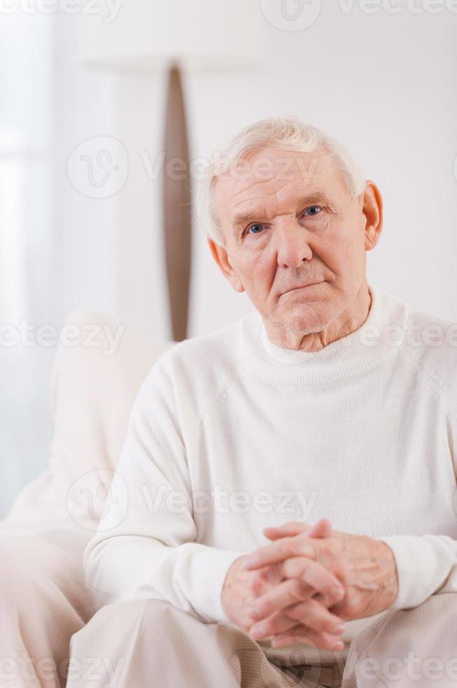 Thoughtful senior man. Serious senior man holding fingers crossed on his hands and looking at camera while sitting in chair photo