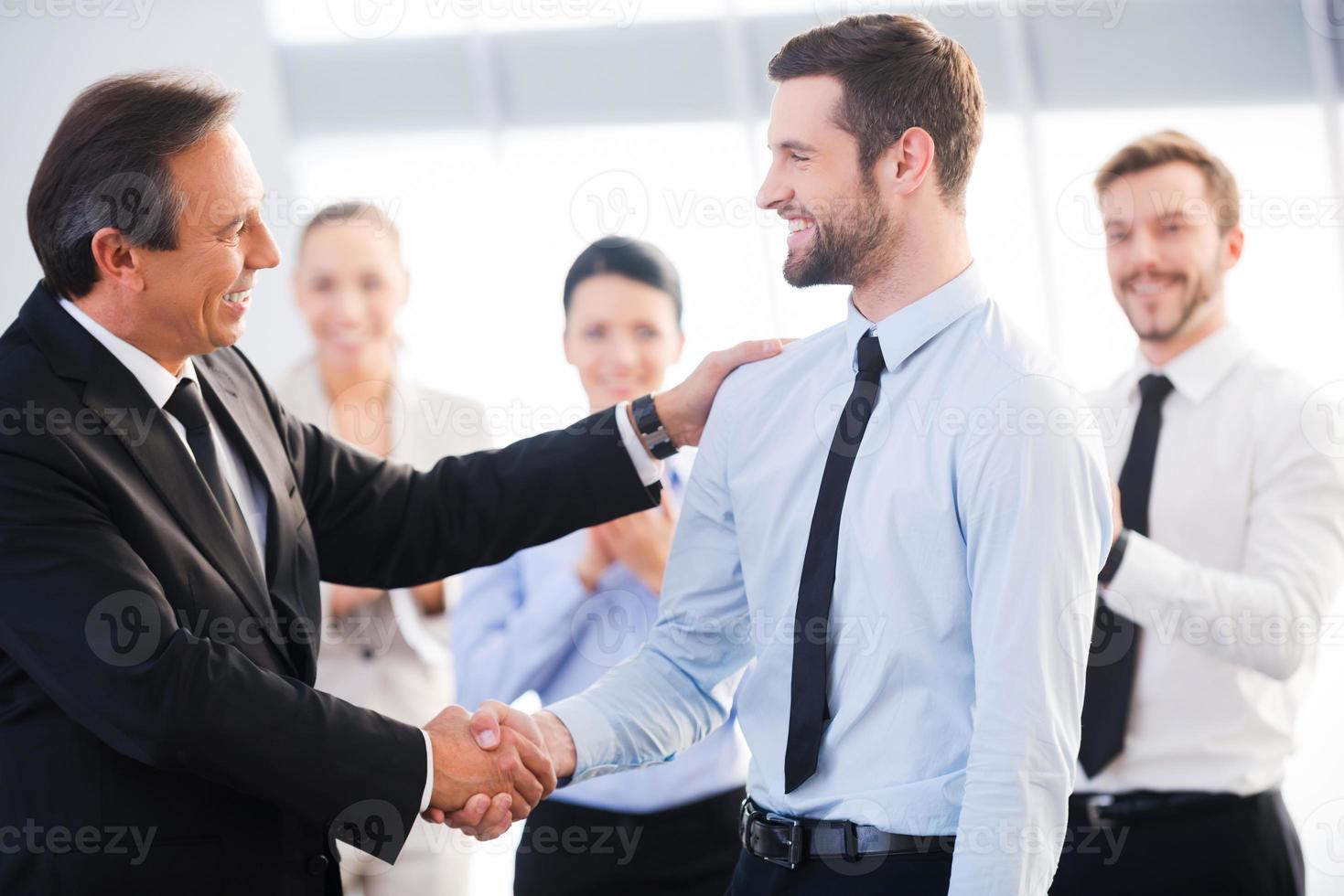 Good job Two cheerful business men shaking hands while their colleagues applauding and smiling in the background photo