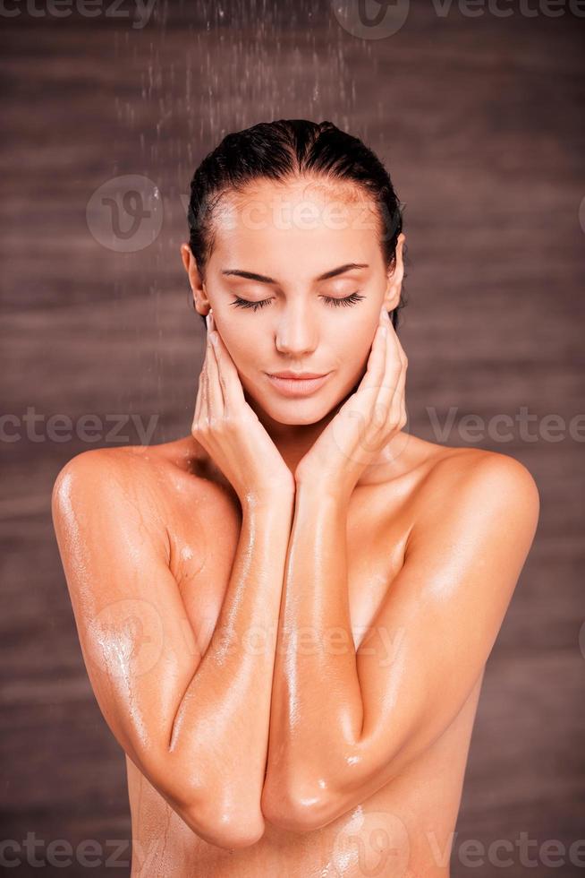 lavando el estrés. hermosa joven sin camisa parada en la ducha y manteniendo los ojos cerrados foto
