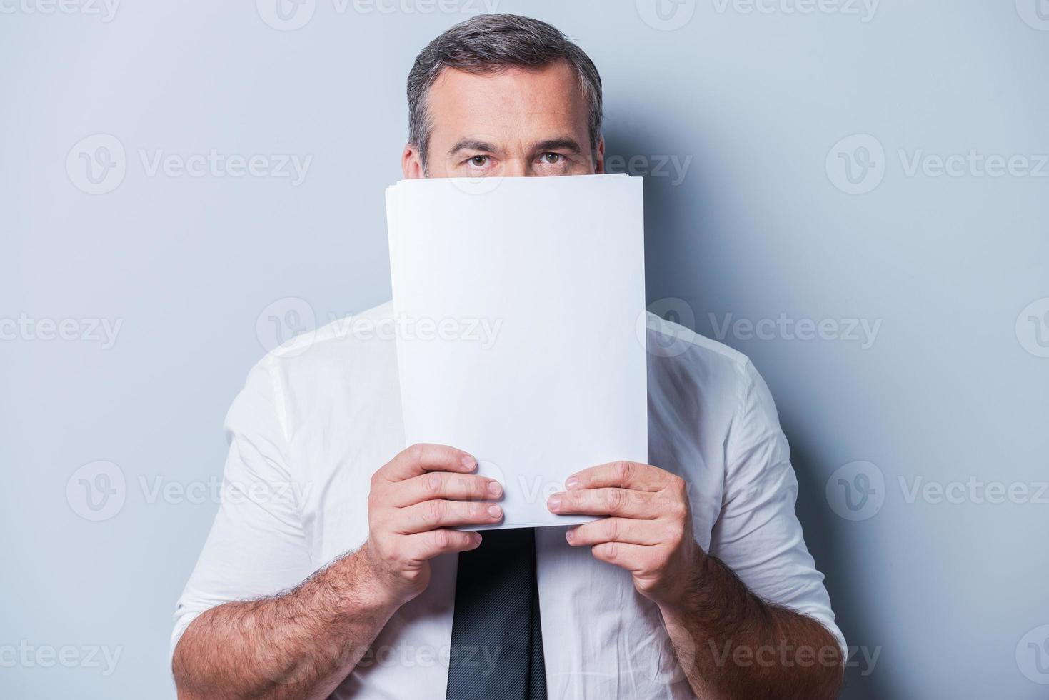 Stuck in paperwork. Confident mature man in formalwear hiding part of his face behind documents and looking at camera while standing against grey background photo