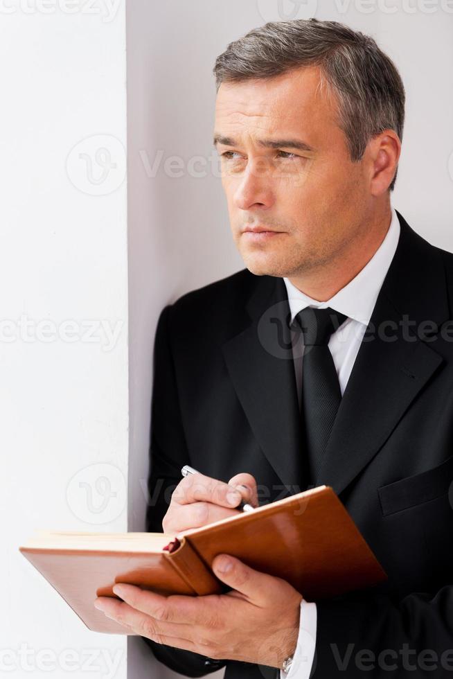 Looking for inspiration. Thoughtful mature man in formalwear writing something in note pad and looking away while leaning at the wall photo