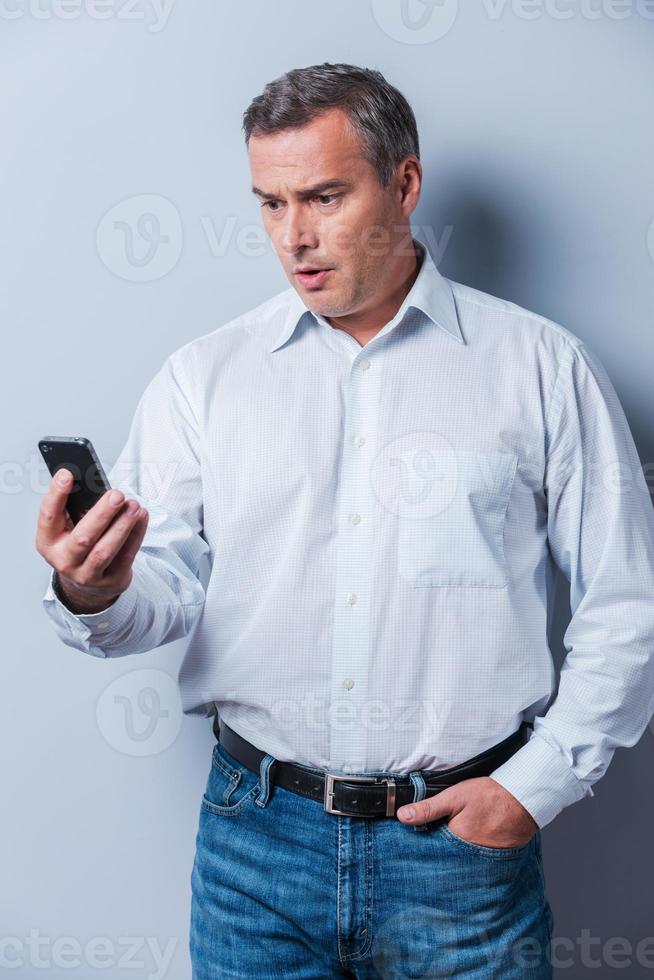Shocking news. Surprised mature man in shirt holding mobile phone and looking at it while standing against grey background photo