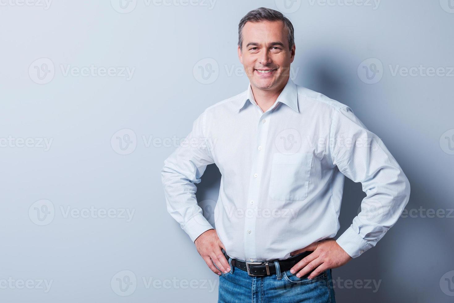 Confident and successful. Confident mature man in shirt looking at camera and smiling while holding hands on hip and standing against grey background photo
