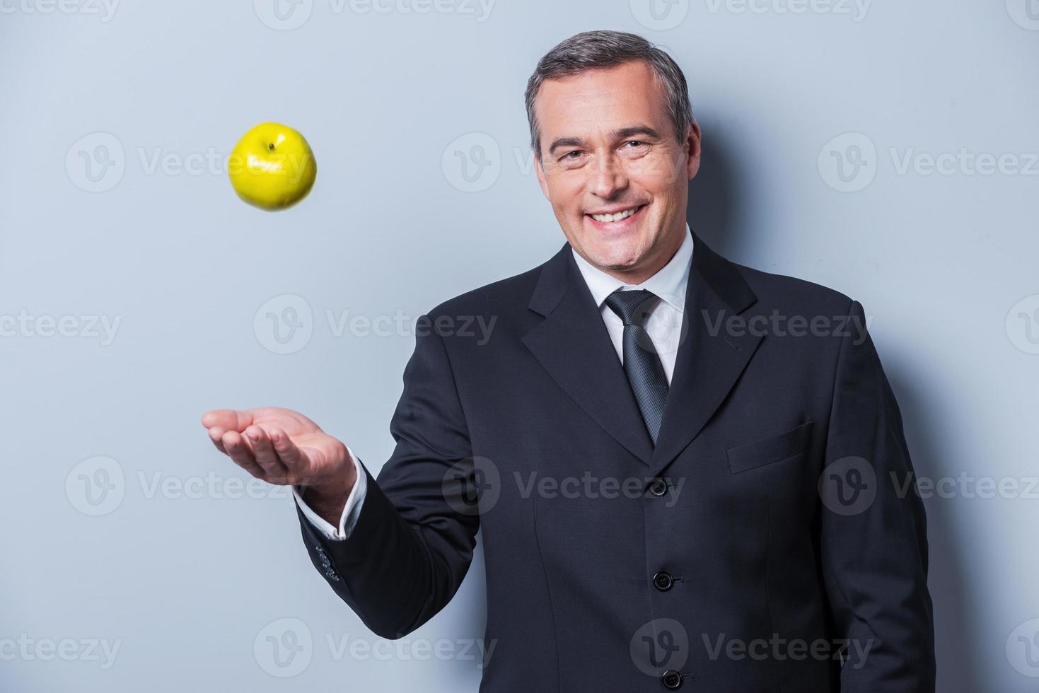 Healthy food for success. Confident mature man in formalwear throwing a green apple up and smiling while standing against grey background photo