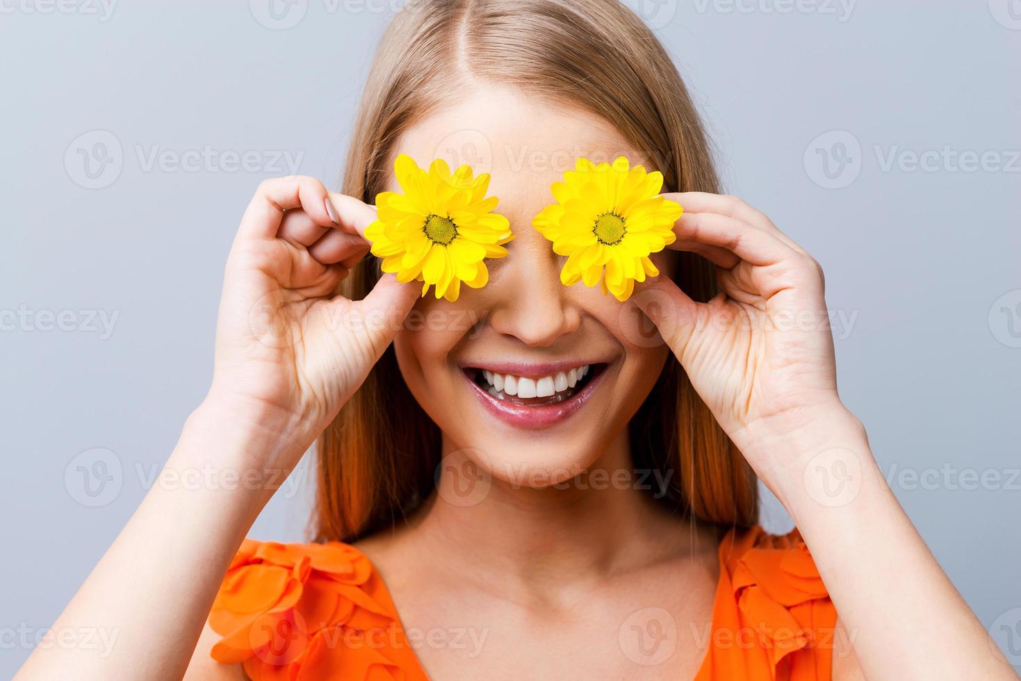 Summer mood. Beautiful young woman in pretty dress holding flowers in front of her eyes while standing against grey background photo