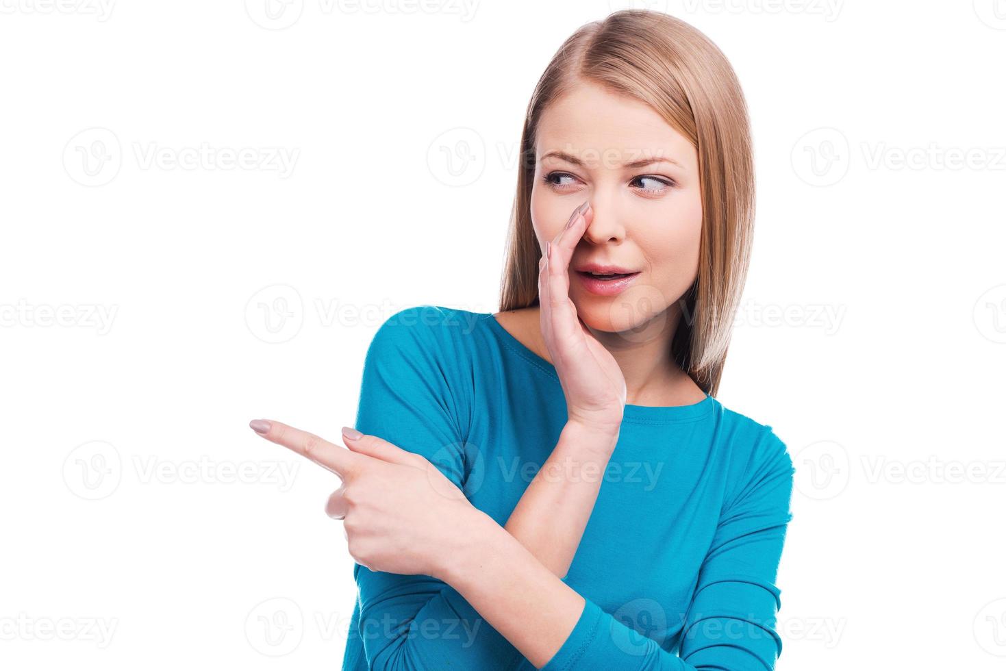 Gossip girl. Beautiful young woman holding hand near mouth and pointing away while standing against white background photo