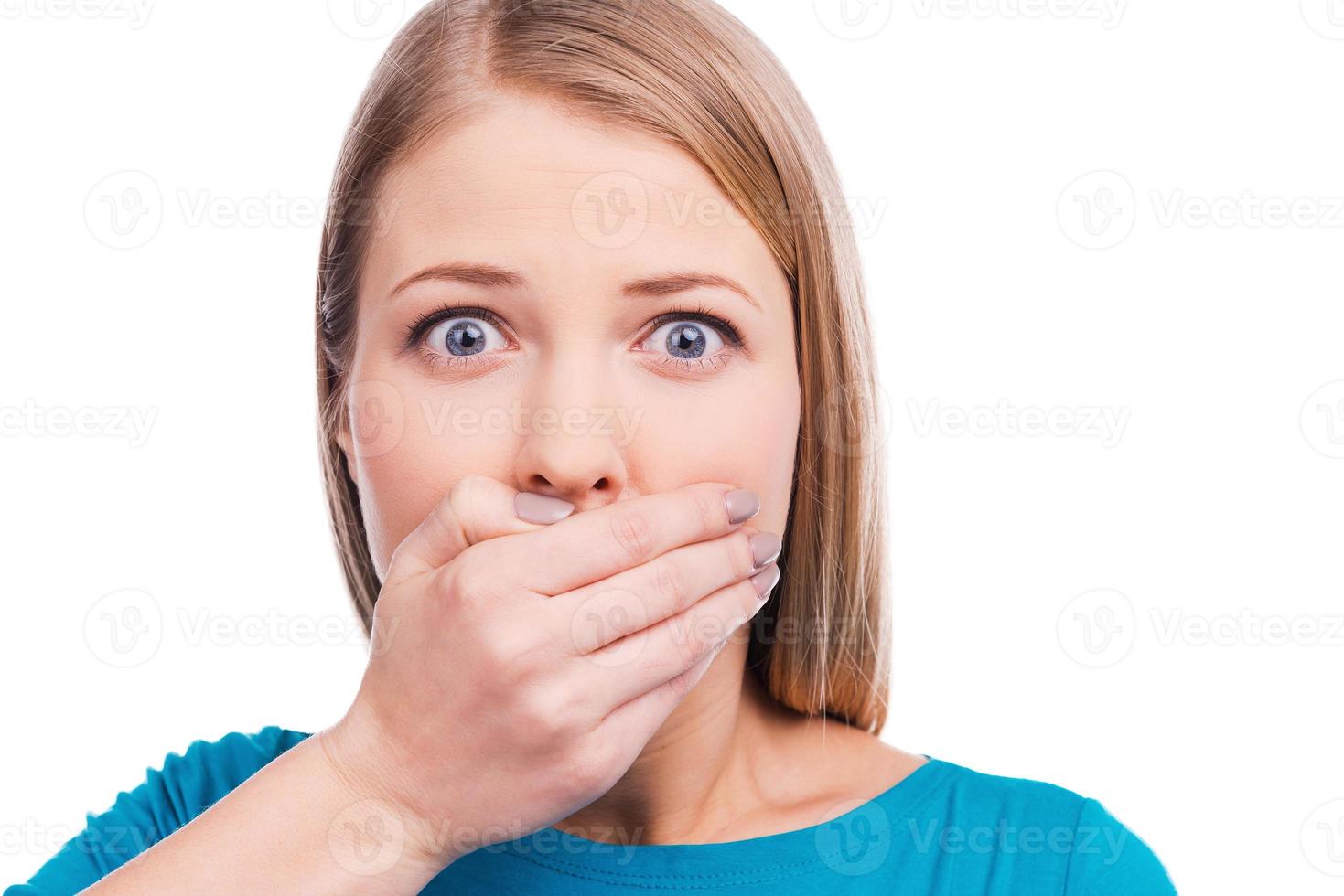 Oops Surprised young woman covering mouth with hand and staring at camera while standing against white background photo