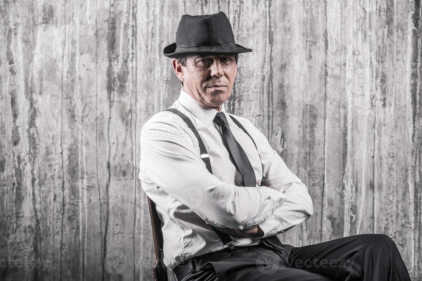 Hidden threat. Bossy senior man in gangster clothing sitting at the chair and looking at camera with a grey wall in the background photo