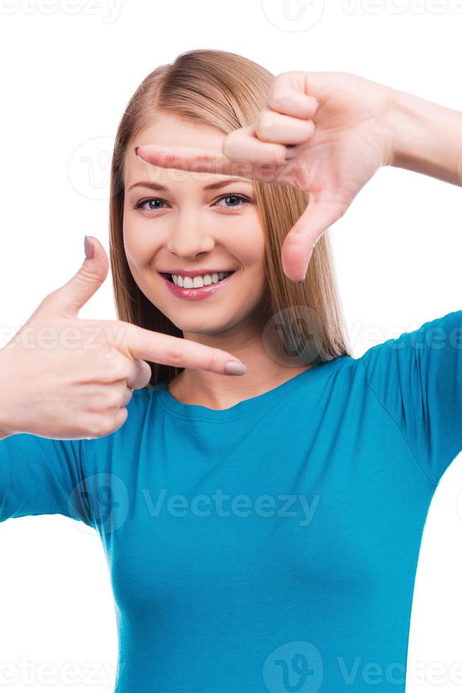 Focusing at you. Beautiful young woman looking at camera and gesturing finger frame while standing against white background photo