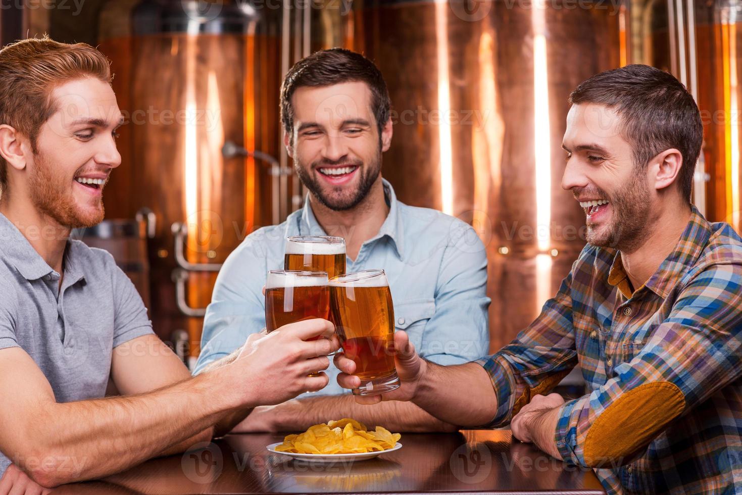 reunirse con viejos amigos en el bar. tres jóvenes felices con ropa informal brindando con cerveza y sonriendo mientras se sientan juntos en un pub de cerveza foto