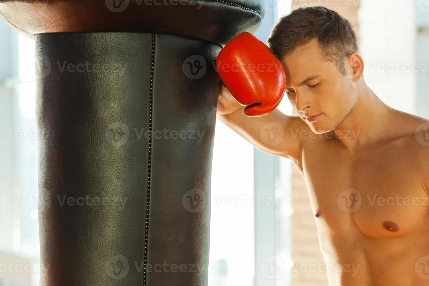 Feeling tired after training. Tired young boxer in sports gloves leaning at the punching bag photo