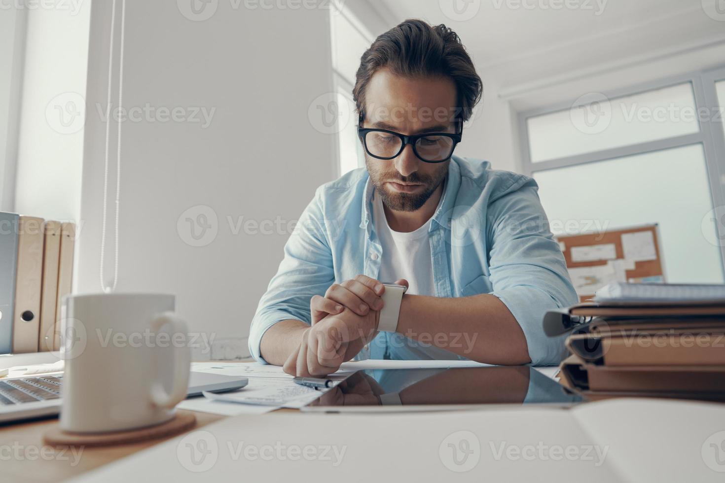 Handsome young man looking at his smart watch while sitting at his working place in office photo