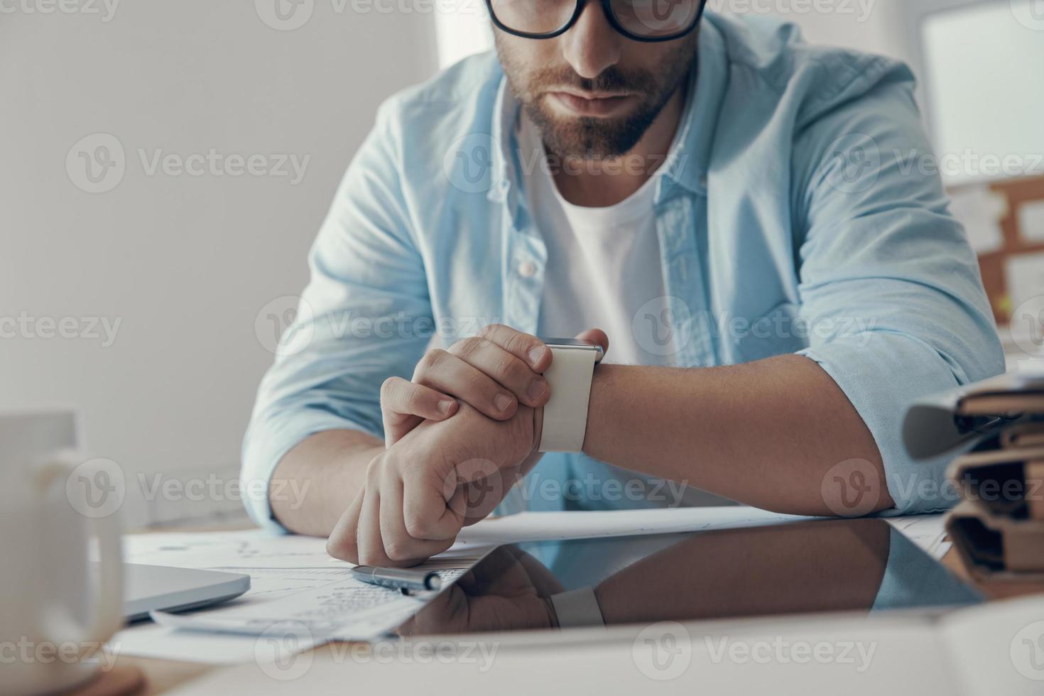 Close-up of young man looking at smart watch while sitting at his working place in office photo