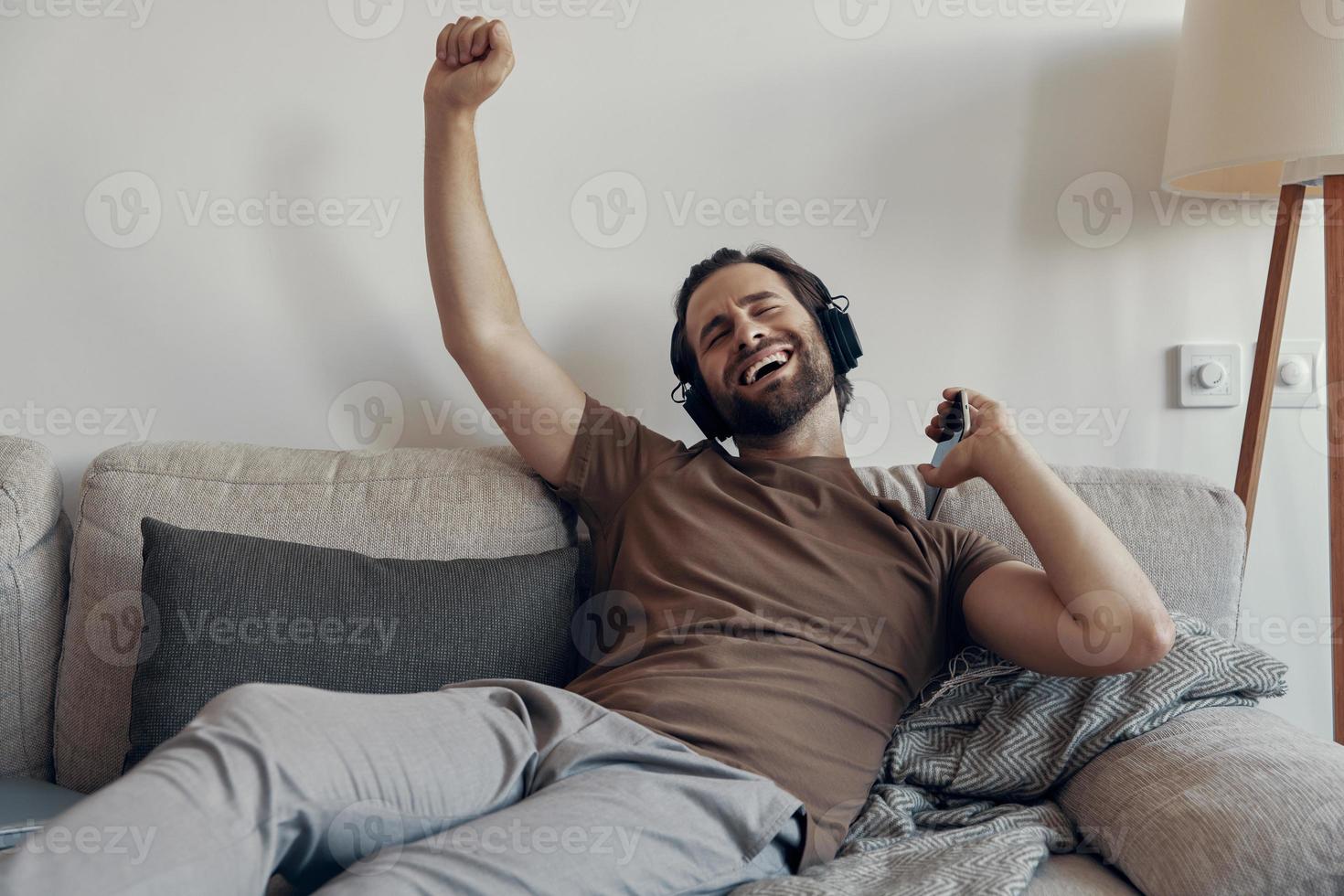 joven feliz con auriculares sosteniendo un teléfono inteligente y gesticulando mientras se relaja en el sofá foto