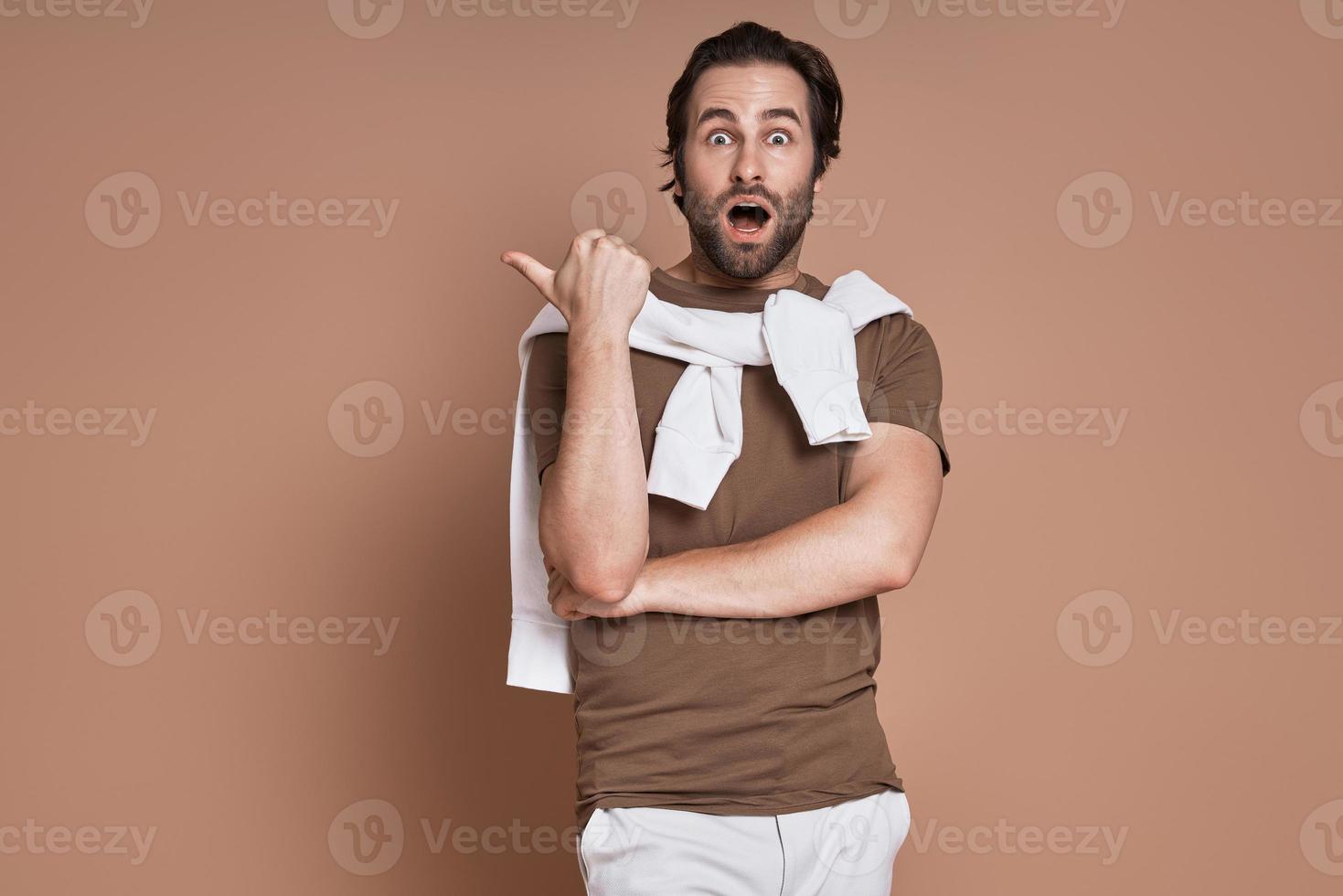 Shocked young man pointing away while standing against brown background photo