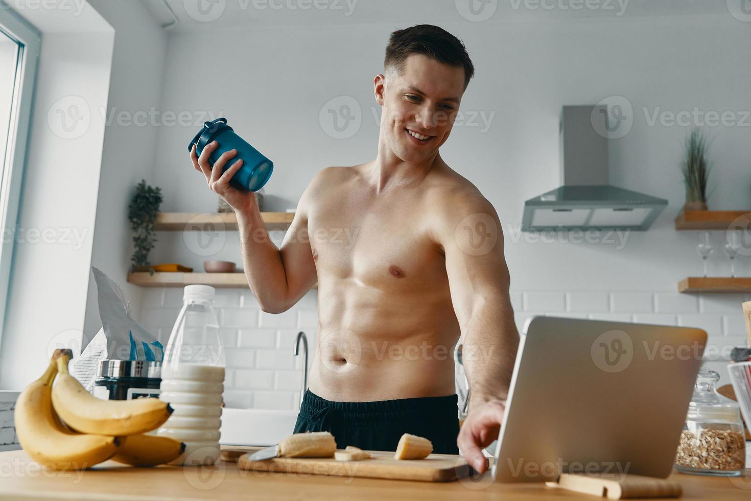 Happy young man preparing protein drink and using digital tablet while standing at the kitchen photo