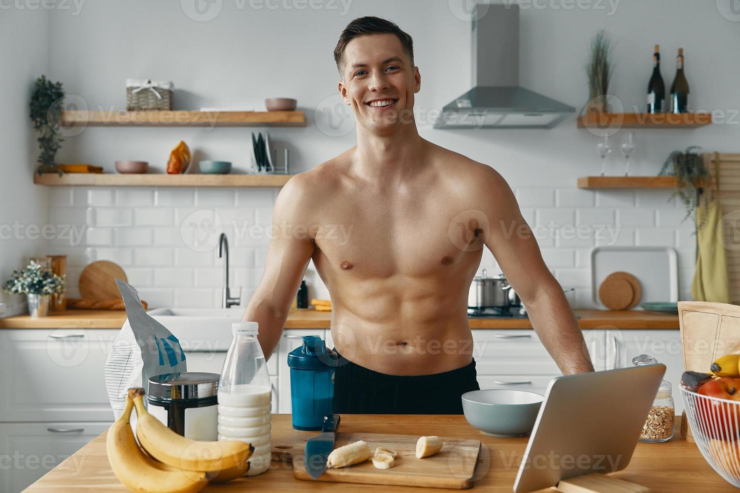 Handsome young man looking at camera and smiling while preparing healthy food at the kitchen photo