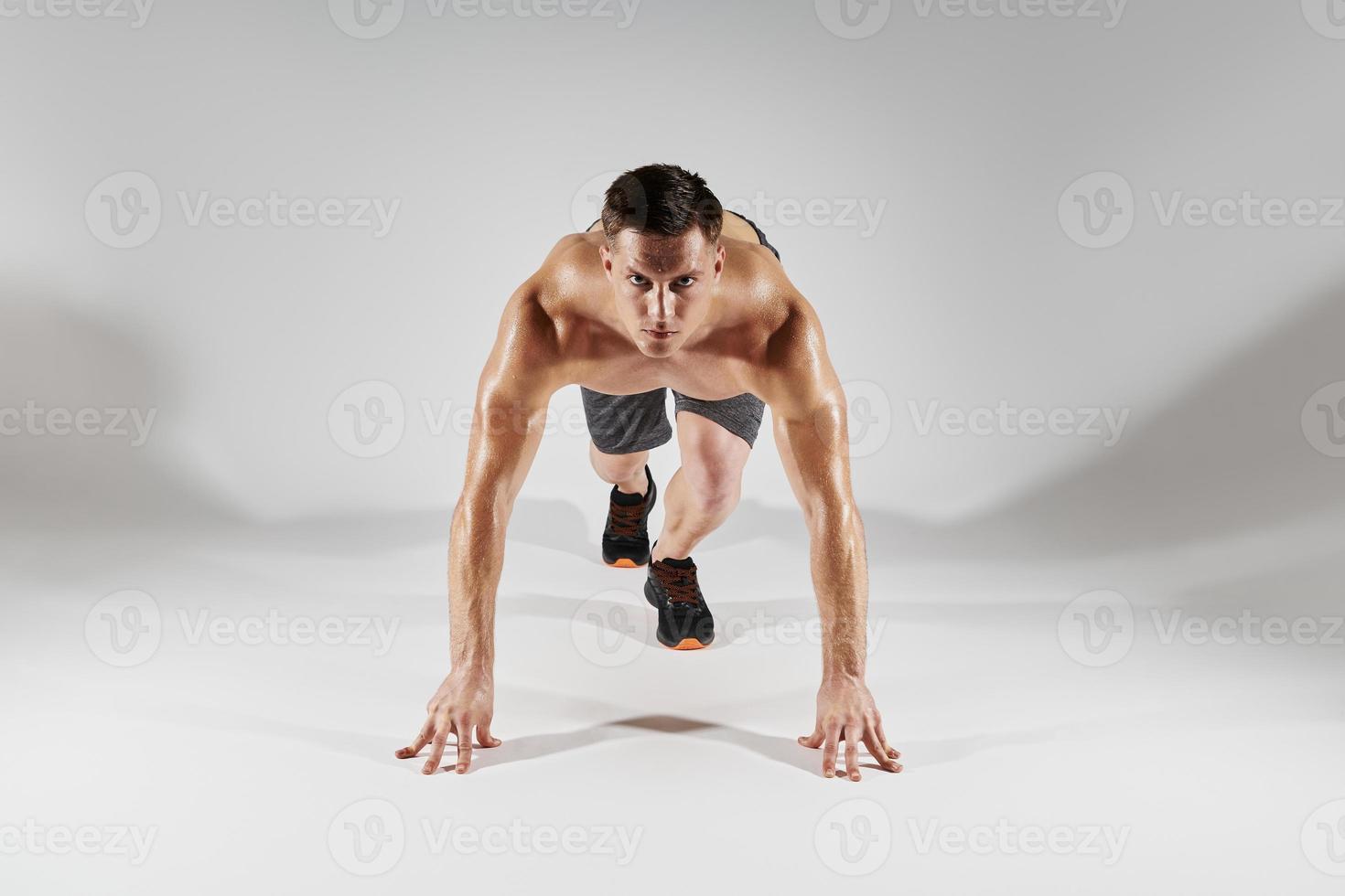 Confident fit man preparing to run while standing in starting position against white background photo