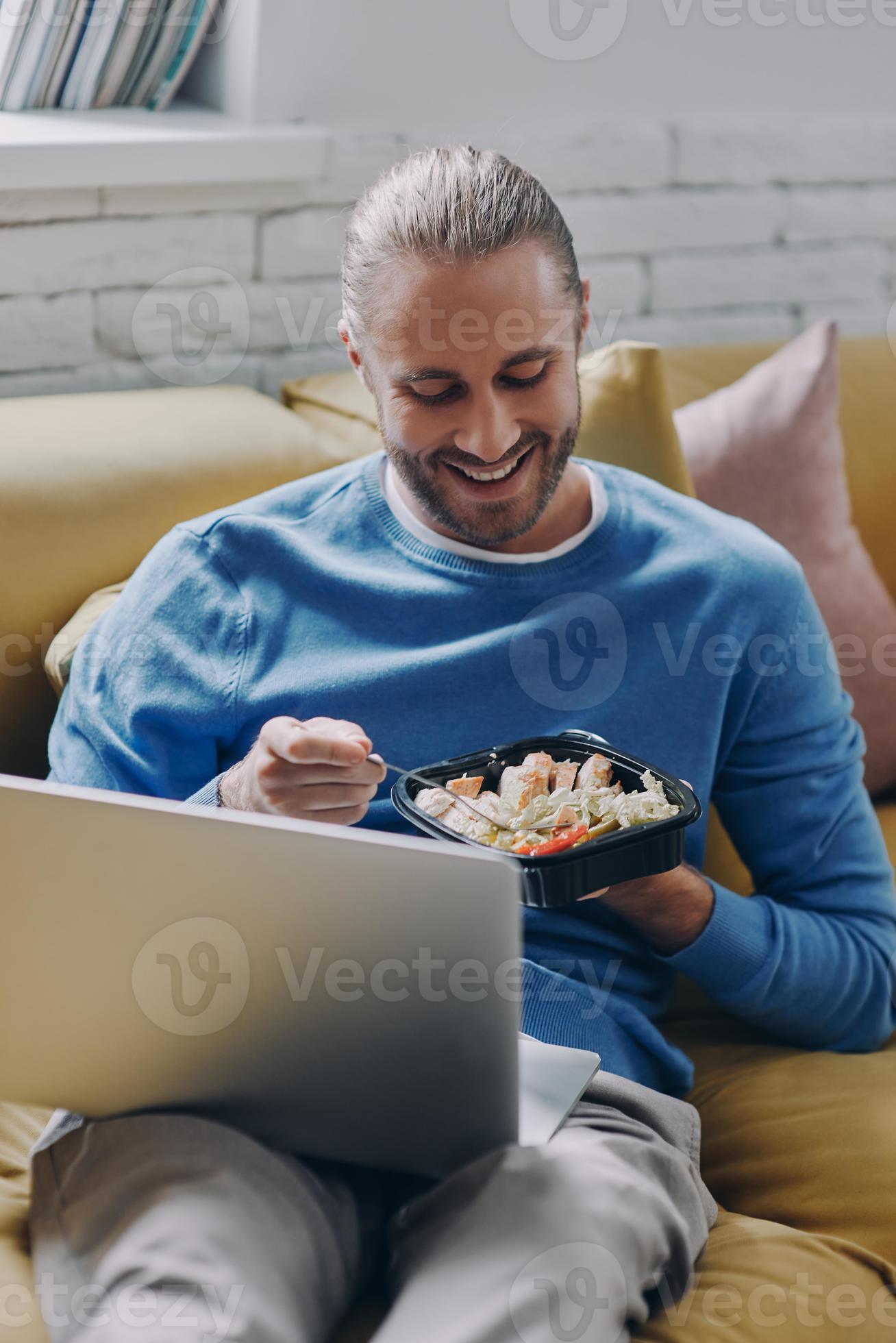 Relaxed young man eating lunch and using laptop while sitting on the