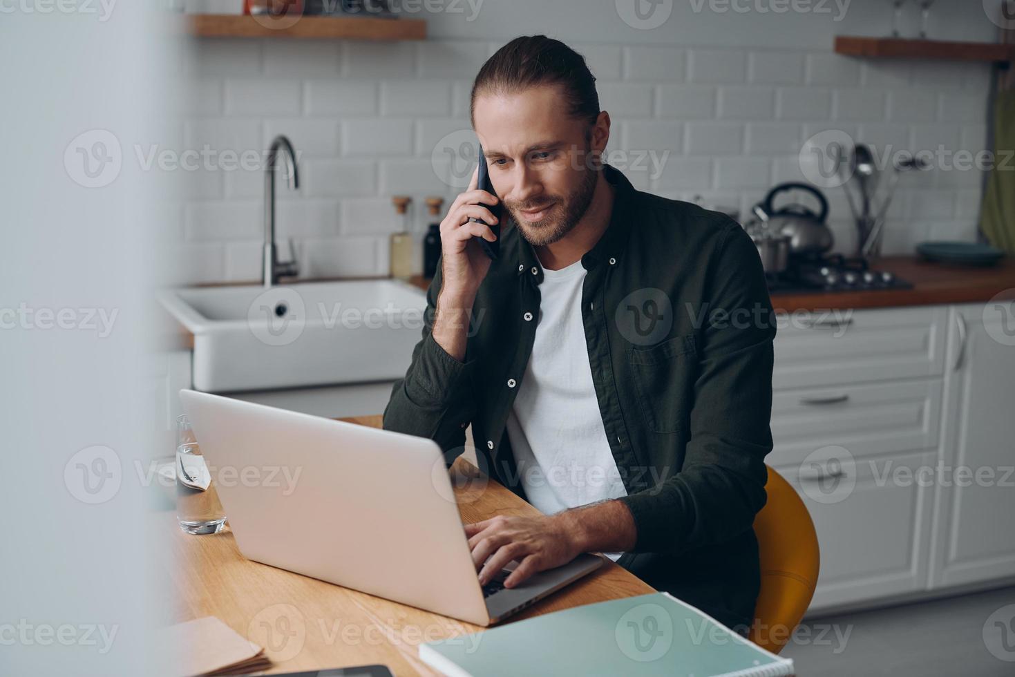 Confident young man talking on mobile phone and using laptop while sitting at the domestic kitchen photo