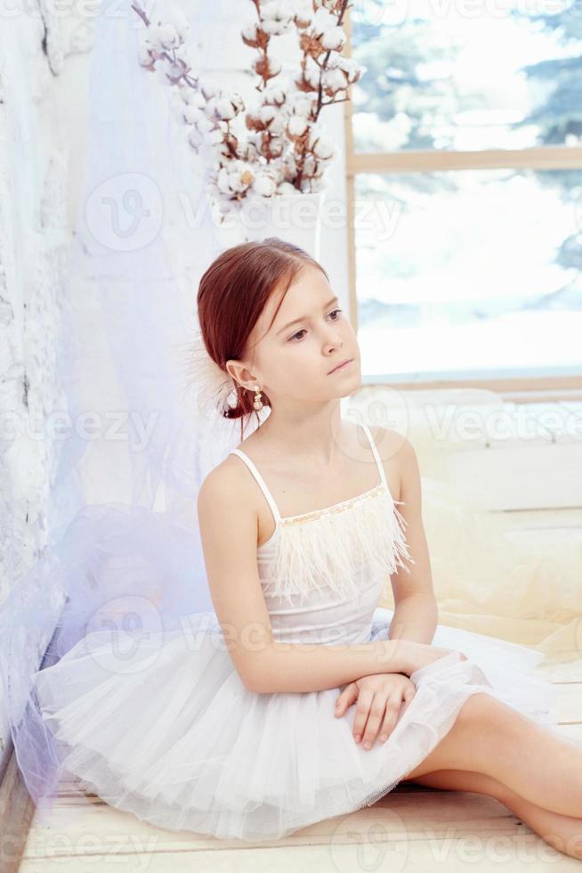 Little prima ballet. Young ballerina girl is preparing for a ballet performance. Girl in a white ball gown and Pointe near the window, beautiful red hair. Young theater actress photo