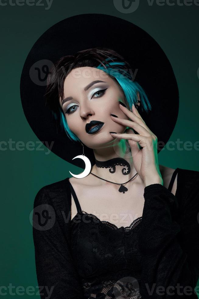 Young Halloween witch in a round hat and green round glasses on a green dark background. Dyed hair, dark lipstick, crescent moon earrings photo