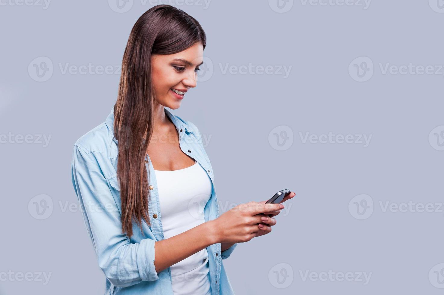 A message from friend.  Smiling beautiful woman chatting with her friends by her smartphone while standing against grey background photo