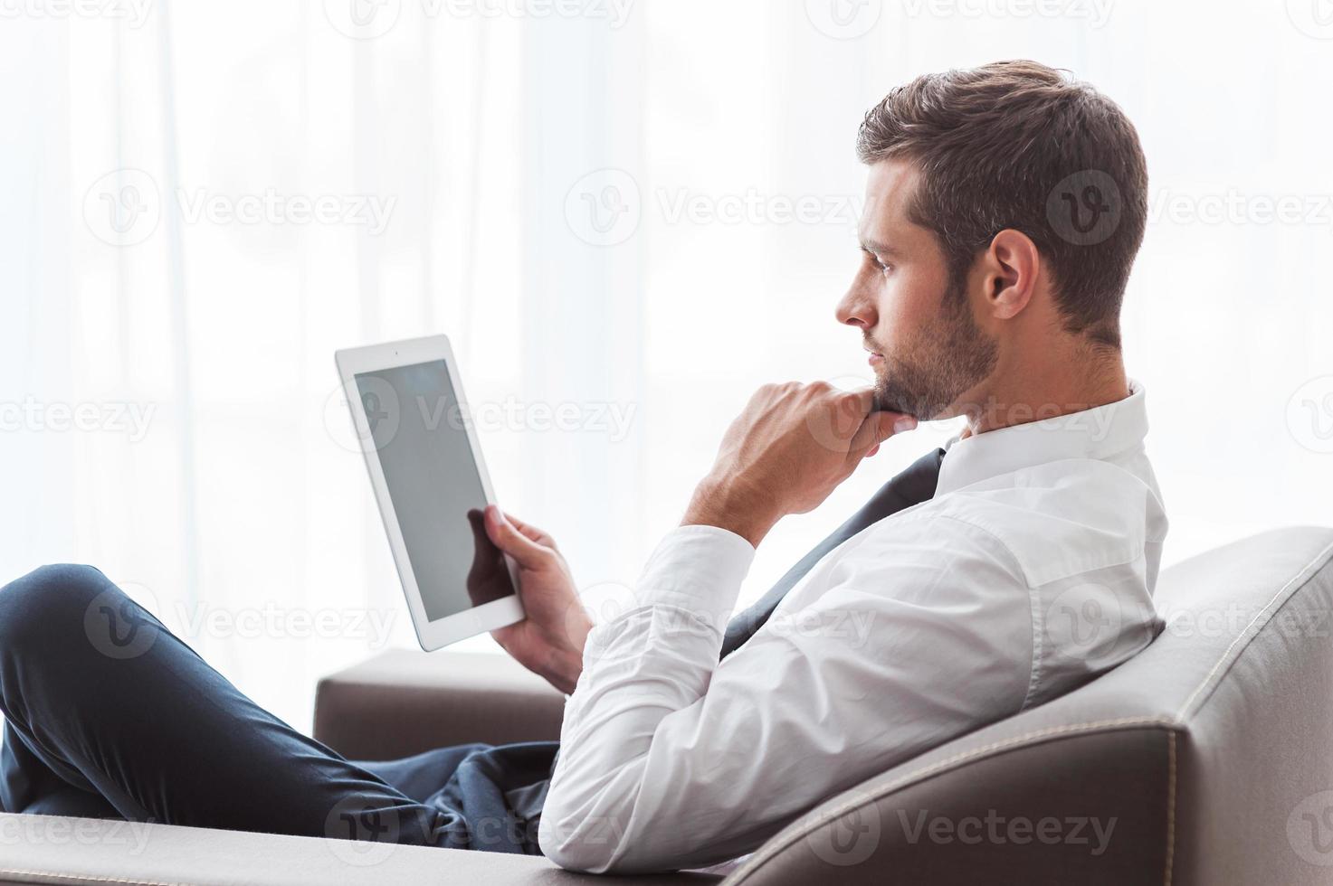Businessman with digital tablet. Side view of confident young businessman in shirt and tie working on digital tablet while sitting at the chair photo