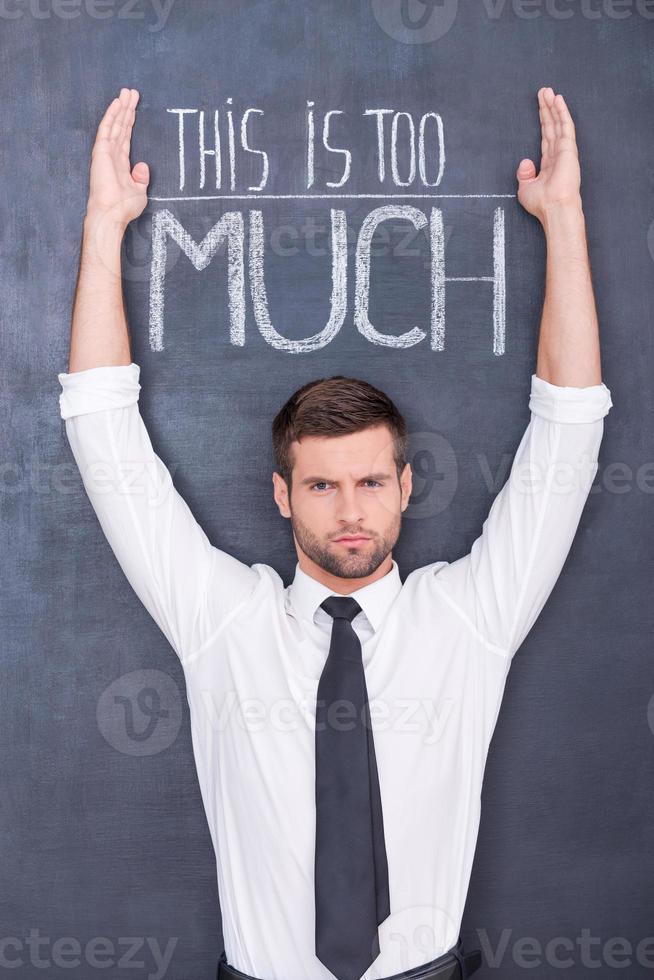This is too much. Confused young man standing against chalkboard with raised hands photo