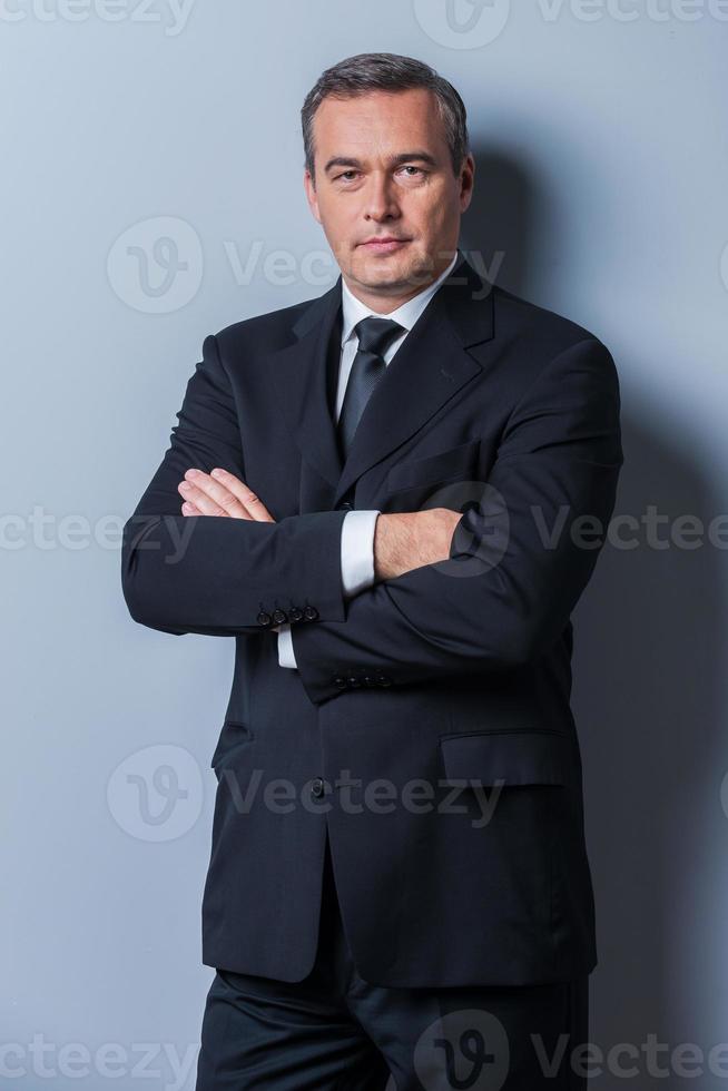 Confident business expert. Confident mature man in formalwear looking at camera and keeping arms crossed while standing against grey background photo