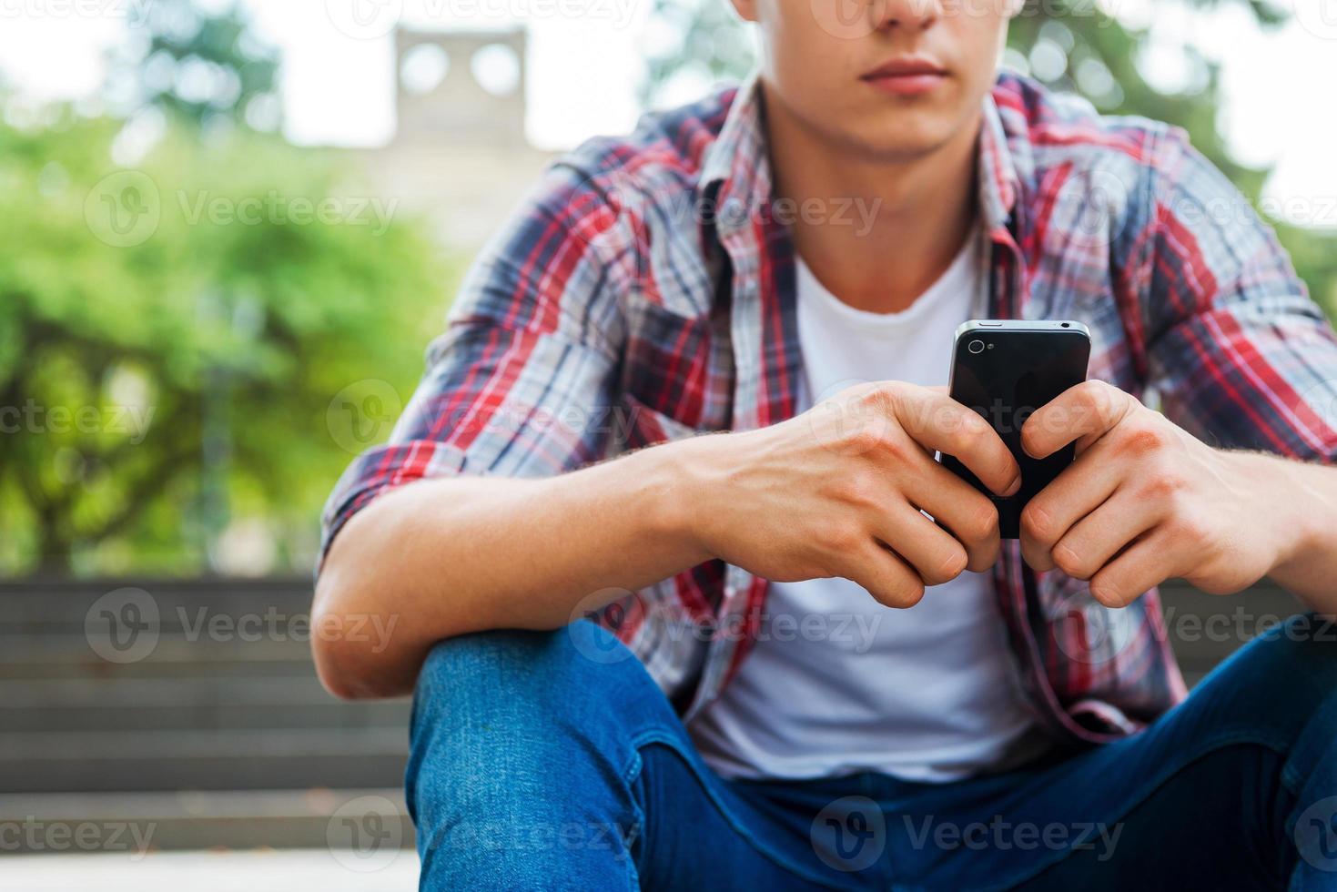 Typing message to friend. Close-up of male student holding mobile phone while sitting at outdoors staircase photo