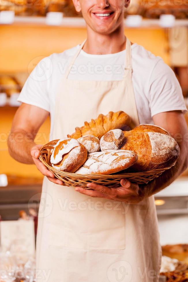 The best baked goods in town. Cropped image of young man in apron holding basket with baked goods and smiling while standing in bakery shop photo