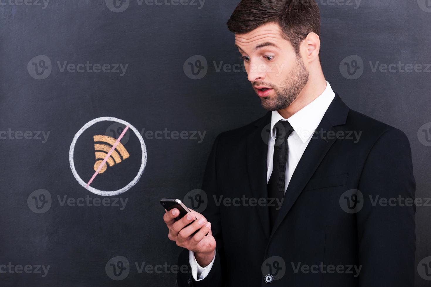 No Wi-Fi. Frustrated young man holding mobile phone and looking surprised because of bad connection while standing against sharing  symbol chalk drawing on blackboard photo