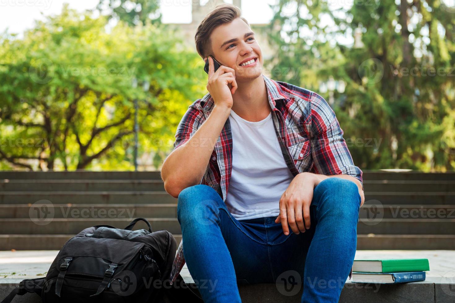 Good news from friend. Happy male student talking on the mobile phone and smiling while sitting at the outdoors staircase with books and backpack laying near him photo