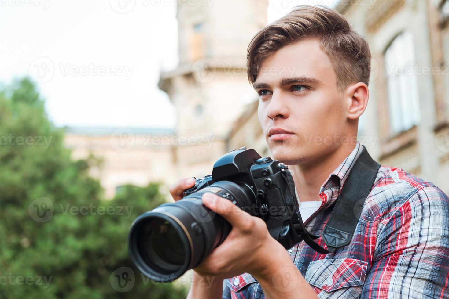 listo para disparar apuesto joven sosteniendo una cámara digital y mirando hacia otro lado mientras está de pie al aire libre foto