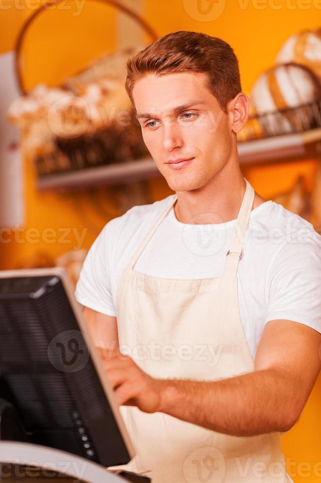 cajero confiado en el trabajo. apuesto joven cajero masculino en delantal trabajando con caja registradora mientras está de pie en la panadería foto